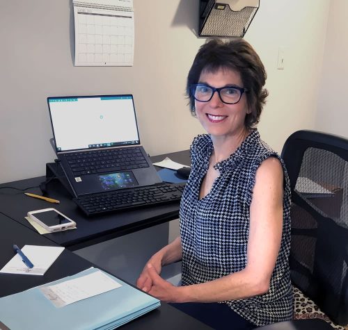 A Woman Sitting At A Desk With A Laptop And Smiling At The Camera